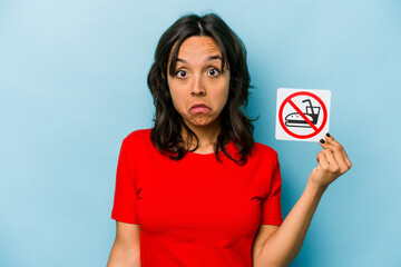 Young hispanic woman holding no eating sign isolated on blue background shrugs shoulders and open eyes confused.