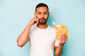 Young hispanic man holding a bag of chips isolated on blue background pointing temple with finger, thinking, focused on a task.