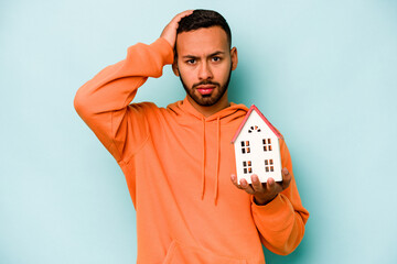 Young hispanic man holding a toy house isolated on blue background being shocked, she has...