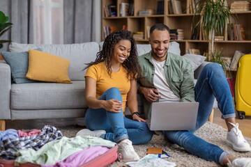 Excited black spouses using laptop while preparing for trip, choosing travel destination and booking tickets online