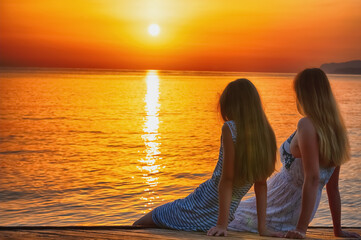 Rear view of mom and daughter by the sea meeting morning sunrise. Summer holiday, rest, communication and travel concept.