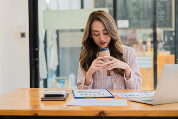 Asian business woman holding a cup of coffee at the desk Financial graph with desktop and laptop calculator for modern office ecommerce marketing ideas.