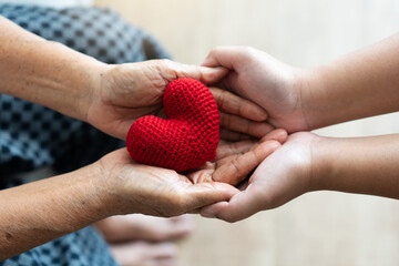 Young and senior woman holding each other hands and red yarn heart shape togetherness concept. Elderly care and protection with love from grandchild.