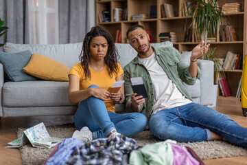 Unhappy african american spouses after tour cancellation, sitting on floor with unpacked clothes, holding travel tickets
