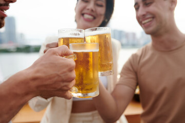 Close-up image of young people clinking mugs with cold beer at brewery bar