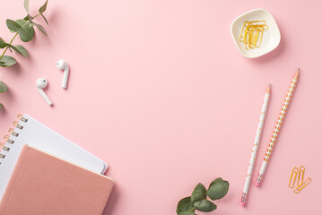 Business concept. Top view photo of workstation pink copybooks wireless earbuds pencils clips and eucalyptus on isolated pink background with empty space