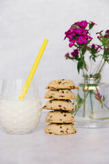 5 chocolate chip cookies are stacked on stop of each other to make a tower. Next to the stack of cookies is a glass of milk and a yellow straw. In the background are purple flowers on a vase. White