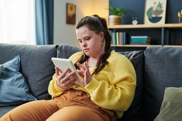 Portrait of modern girl with Down syndrome sitting on couch in living room at home reading book on digital tablet
