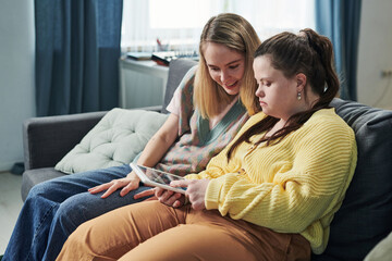 Modern young woman with Down syndrome spending time with her sister at home watching content in social net on digital tablet