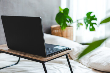 Setup of laptop on portable table with coffee and tea mug and sticky notes on bed with stack of documents and papers with graphs and charts placed on clean white sheet on bed in room