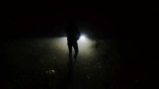 Dark male figure going through forest or countryside in light of car headlights at night. Search for missing person late at evening in natural area. Faceless man silhouette looking for someone