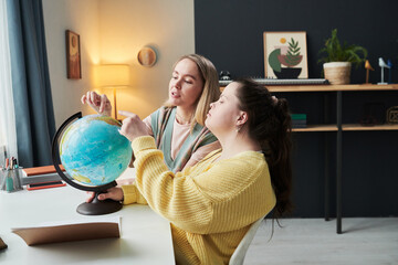 Young girl with Down syndrome sitting at desk next to her teacher turning globe and talking about countries and cultures