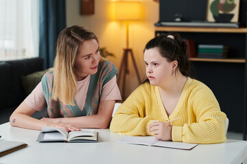 Young Caucasian woman working as teacher sitting at table next to female student with Down syndrome...