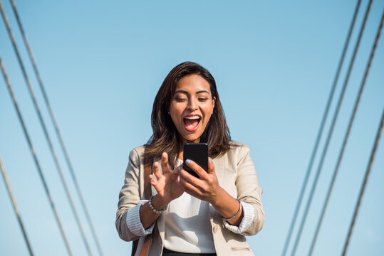 Portrait Of A Young Latina Excitedly Looking At Her Cell Phone, Smart Phone On The Street