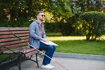 Blind man reading braille book, sitting on bench in summer park, resting