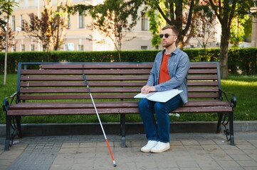 Visually impaired man with walking stick, sitting on bench in city park. Copy space