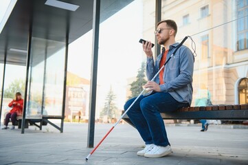 Young blind man with smartphone sitting on bench in park in city, calling. - obrazy, fototapety, plakaty