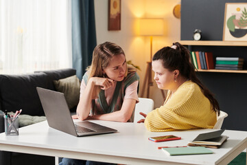 Young woman with Down syndrome and her university friend sitting at table working together on project looking at laptop screen and discussing issue at home