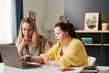 Pensive Caucasian woman and her co-worker with Down syndrome sitting in front of laptop working on...