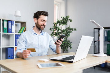 Fototapeta premium Successful online shopping through a smartphone. Successful deal. Satisfied young man holds a credit card and a mobile phone, makes an order. Sitting at a desk in the office on a laptop