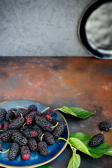 Vertical shot of fresh mulberry with leaves on blue ceramic plate on grunge rusty metal surface and black plate on background. Close-up. Selective focus, blurred background.