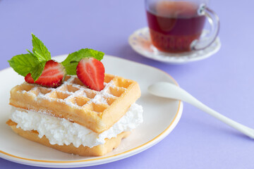 Delicious belgian waffles with whipped cream, strawberry and mint. Waffle sandwich on a plate with dessert spoon and cup of tea on lilac background. Close-up. Selective focus, blurred background.