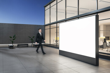 Fototapeta premium Side view businessman passing by outdoor blank white billboard and stylish street benches near modern business center building with glass walls in the evening