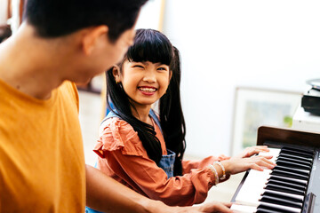 Cheerful Asian daughter in casual clothes with pigtails smiling and looking at dad while leaning to play piano in daytime at home - 506496909