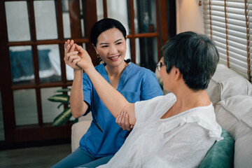 Delightful female caregiver in blue uniform smiling and stretching arm of elderly Asian woman while sitting on sofa against window in living room at home