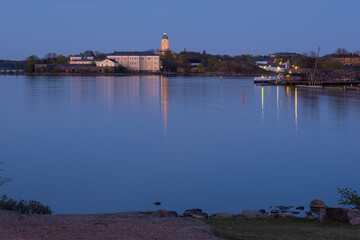 Suomenlinna fortress across the river. A lighthouse casting reflections on the calm sea.
