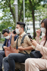 Frowning young Asian man in protective mask talking on phone when sitting on bench outdoors