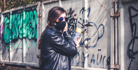 A girl in a black jacket adjusts her mask, holding a spray can of paint in her hand