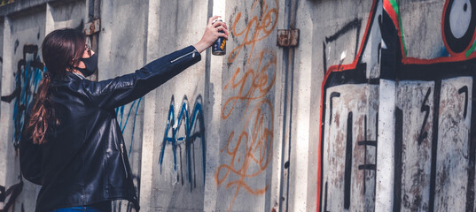 A girl in a black mask and black jacket draws graffiti on the wall in the park
