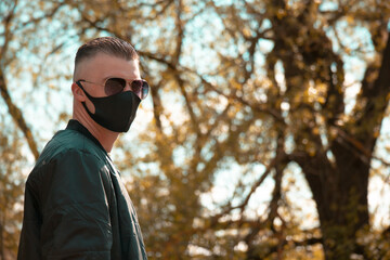 Fototapeta na wymiar A guy in glasses and a protective black mask looks over his shoulder on a sunny day. The guy is short-shaven, in a black jacket against the background of trees. Photo of excellent quality