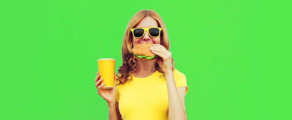 Portrait of happy smiling young woman with fast food, burger and cup of juice wearing yellow t-shirt, sunglasses on green background