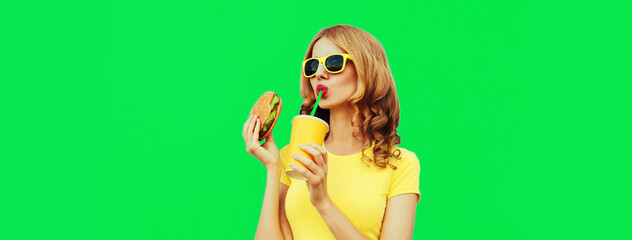 Portrait of young woman drinking fresh juice with fast food, burger wearing yellow t-shirt, sunglasses on green background