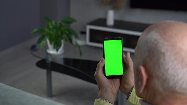 Older grey haired man is sitting on sofa at living room and swiping phone with blank green screen with mockup