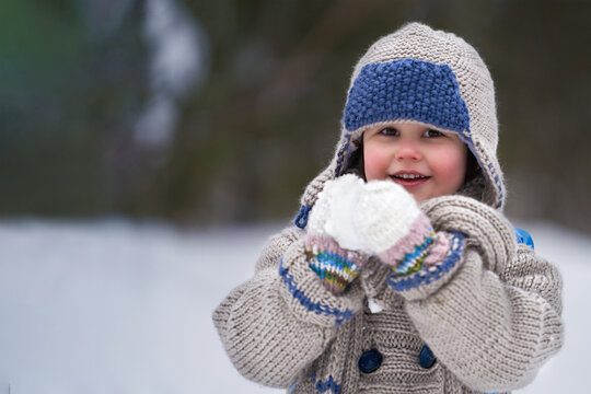  A happy little girl with a snowball in her hands on a blurry background of a winter forest. Copy space. Selective focus.