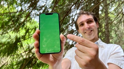 very handsome man french italy not holding a phone with a green tap standing in the woods in a white shirt and smiling showing a finger on the screen can use for advertising. High quality 4k footage