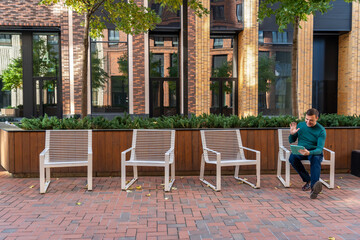 Millennial man with happy face greets friends or colleagues on tablet phone screen, sitting on one of four chairs in courtyard of modern residential complex with landscaping. Portrait of excited man