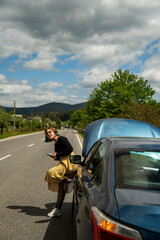 Caucasian hipster girl using a mobile phone to call the mechanic while looking at broken down car on street. Young girl with a broken car on the road call service. Cute girl near a broken car