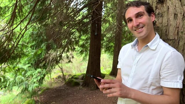 very handsome man french italy not holding a phone with a green tap standing in the woods in a white shirt and smiling showing a finger on the screen can use for advertising. High quality 4k footage