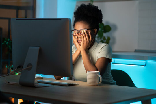 Symptoms of overworking. Young tired overworked african woman freelancer in eyeglasses working late from home, sitting in front of monitor, feeling eye strain and fatigue during computer work. 