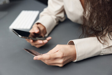 Cool Mockup offer. Side of view on hands of tanned curly Latin woman in linen shirt with credit debit card and modern smartphone on gray background. Copy space. Shopaholic lady doing online shopping.