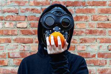 A young girl in a black helmet, glasses and a mask holds an orange to her head.