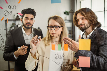 Caucasian female marketer drawing progress graph on transparent wipe board while working with multietnic male colleagues at corporate office. Concept of projects planning and business presentation.
