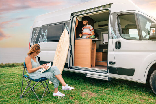Young man cooking vegetables in a camper van while young woman reads outdoors