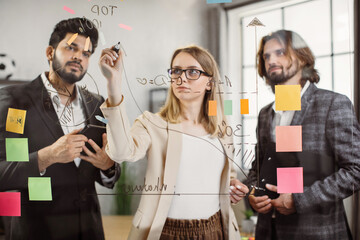 Multicultural group of young financiers in formal wear using transparent glass board during office meeting. Female worker explaining new strategy to male colleagues in boardroom.