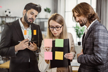 Group of multicultural professional brokers sticking notes with project information on glass wall during office meeting. Partners with tablet and clipboard cooperating together at modern workplace.