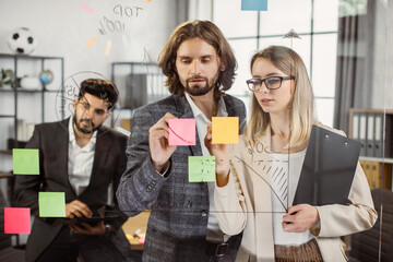 Couple of workers standing in front of glass wall and writing ideas on sticky notes with indian male colleague on background. Young employees in formal wear sharing experience during office meeting.